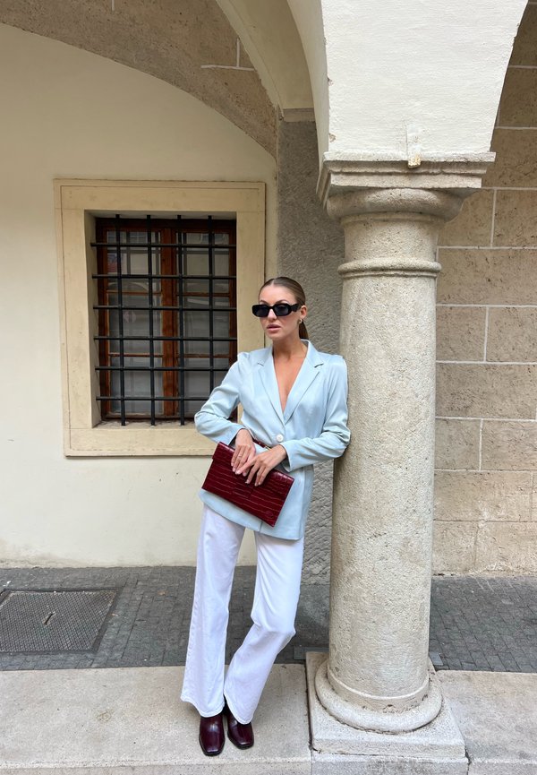 Mujer con blazer azul claro, pantalones blancos, gafas de sol oscuras y sosteniendo un bolso de mano marrón rojizo, apoyada en una columna de piedra al aire libre.