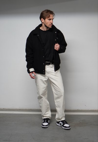 Chaqueta negra con botones a presión sobre una camiseta negra, combinada con pantalones blancos de corte holgado y zapatillas deportivas en blanco y negro. Diseño sencillo y moderno.