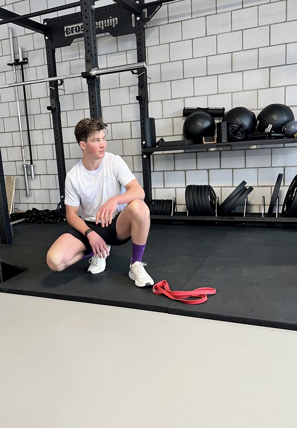 Joven con ropa de entrenamiento descansando sobre una rodilla en el suelo del gimnasio cerca de pesas, una banda de resistencia y balones medicinales en estantes.