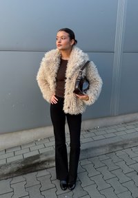 Jakna iz kremaste umetne dlake s puhasto teksturo, temno rjavo oprijeto majico, črne razširjene hlače in sijoči črni čevlji, drži rjavo torbico.