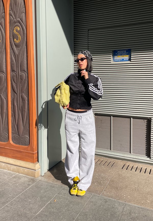 Frau in schwarzer Sportjacke, weißen Jogginghosen, gelben Turnschuhen und Bandana, die eine gelbe Tasche hält und neben einem Gebäude mit Metallrollladen steht.