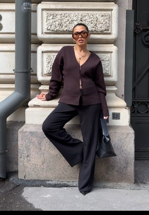 Mujer con gafas de sol grandes, cárdigan oscuro, pantalones anchos negros y tacones, apoyada contra una pared de piedra con textura cerca de una tubería y una puerta.