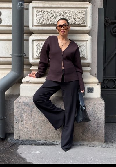 Mujer con gafas de sol grandes, cárdigan oscuro, pantalones anchos negros y tacones, apoyada contra una pared de piedra con textura cerca de una tubería y una puerta.