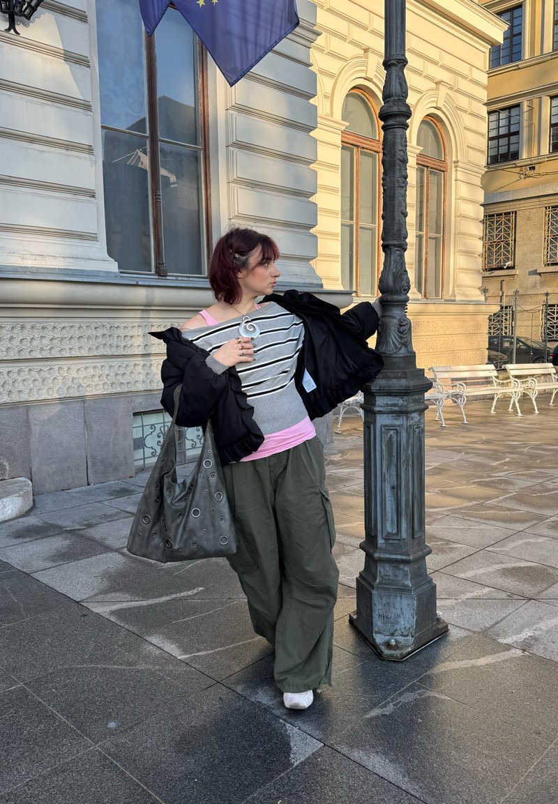 Personne portant une veste noire, un haut rayé gris et blanc, et un pantalon olive, tenant un sac gris avec des découpes circulaires, debout près d'un lampadaire.