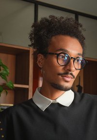 Person med lockigt hår som bär runda svarta glasögon, en svart topp och en vit krage. Trähyllor och en växt i bakgrunden.