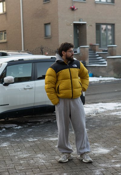 Chaqueta acolchada con cuerpo amarillo y sección de hombros negra, sudadera con capucha gris debajo, pantalones deportivos grises y zapatillas de color claro, de pie al aire libre.