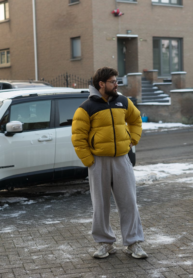 Chaqueta acolchada con cuerpo amarillo y sección de hombros negra, sudadera con capucha gris debajo, pantalones deportivos grises y zapatillas de color claro, de pie al aire libre.