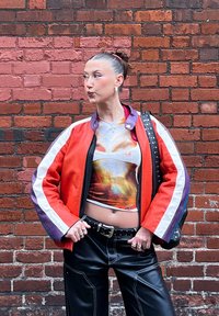 Chaqueta bomber roja y morada con rayas blancas, top corto estampado, pantalones de cuero negros y bolso negro con tachuelas frente a una pared de ladrillo.