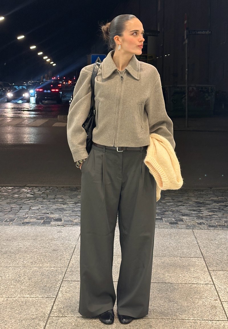 Donna con i capelli raccolti in uno chignon, indossa una giacca beige con cerniera e pantaloni ampi scuri, tiene in mano un maglione crema, in piedi in una strada della città di notte.