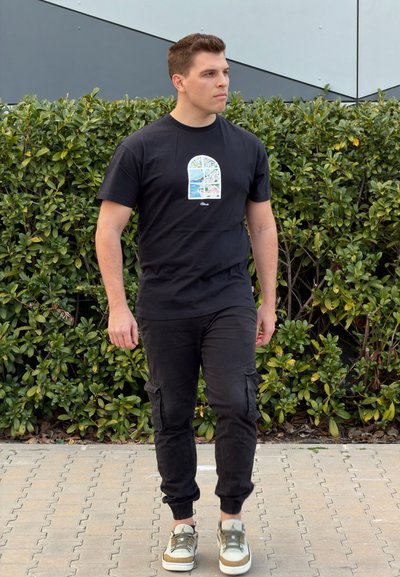 Hombre caminando al aire libre con una camiseta negra con estampado, pantalones cargo negros y zapatillas blancas con detalles verdes.