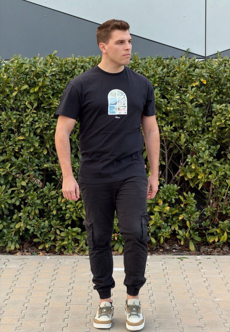 Vīrietis staigā ārā, valkājot melnu grafisko t-kreklu, melnas darba bikses un baltas sporta kurpes ar zaļiem akcentiem.