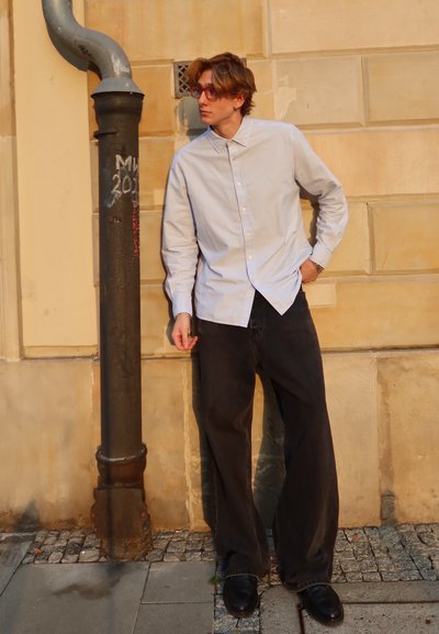 Camisa de botones de manga larga azul claro, pantalones vaqueros negros holgados y zapatos negros. Apoyado contra un poste de metal negro junto a una pared texturizada.