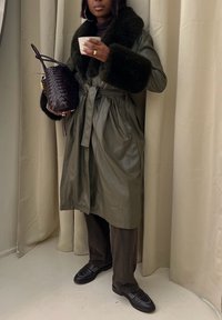 Olivengrüner Leder-Trenchcoat mit einem Kragen aus Kunstfell, tailliertem Gürtel und gefältetem Design. Kombiniert mit dunkelbraunen Hosen und Mokassins.