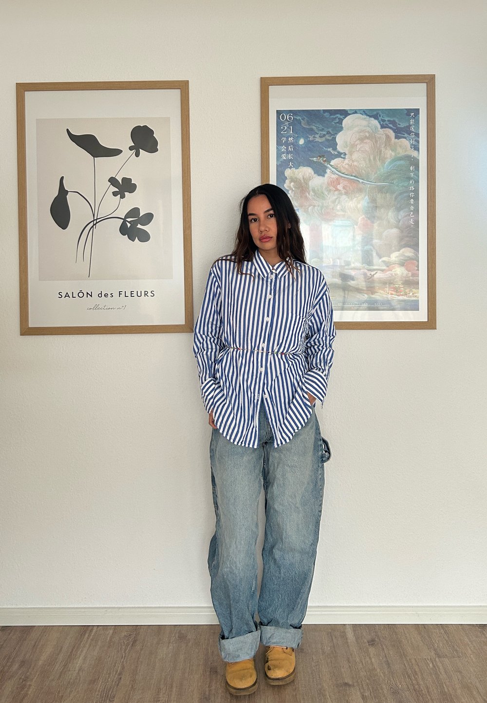 Blå og hvid stribet skjorte med knapper og en frontlomme, parret med løstsiddende lyseblå jeans og brune træsko. Rammer med kunst på væggene.