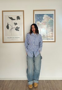Blå og hvid stribet skjorte med knapper og en frontlomme, parret med løstsiddende lyseblå jeans og brune træsko. Rammer med kunst på væggene.