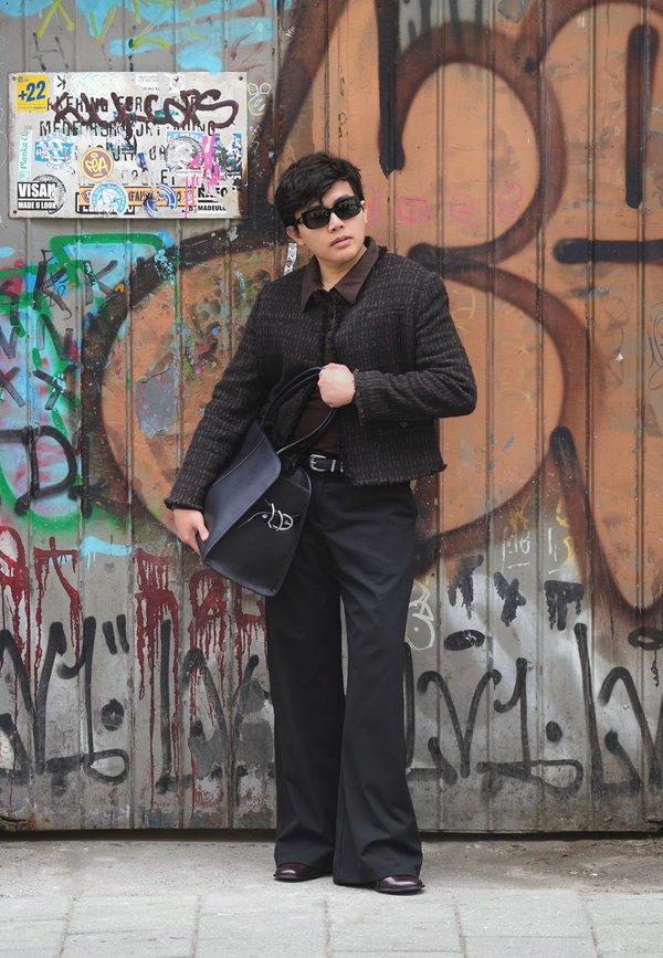 Una persona con gafas de sol y ropa oscura sostiene un bolso negro grande y está de pie frente a una pared metálica cubierta de grafitis.
