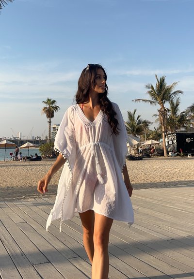 Cubre baño de algodón blanco, de corte holgado, con ribete de pompones y textura punteada; llevado sobre un traje de baño rojo, caminando por un paseo de madera.