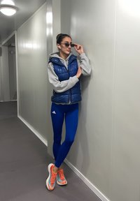 Blauwe pufferbodywarmer over een grijze hoodie, blauwe legging en oranje sportschoenen. Accessoires zijn onder andere een zonnebril en draadloze oordopjes.