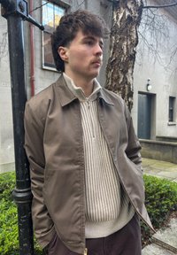 Rjava jakna na zadrgo preko svetle teksturirane jopice z navpičnimi črtami. Model stoji ob teksturirani steni in drevesu.