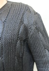 Tumma neulottu pusero, jossa on teksturoitu palmikkokuvio, syvä V-pääntie ja pitkät hihat. Kangas vaikuttaa kestävältä ja joustavalta.