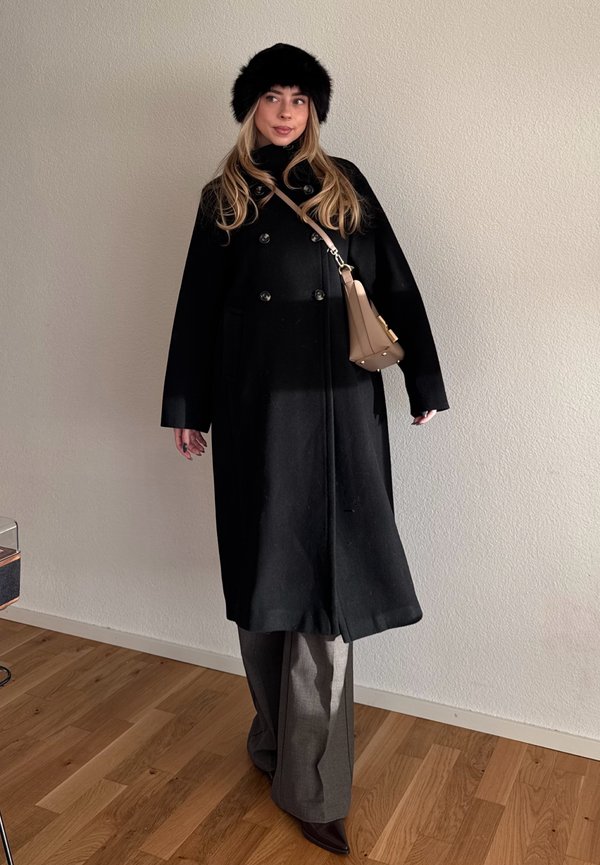 Vrouw met een zwarte bontmuts, lange zwarte jas, grijze wijde broek, bruine puntige schoenen en een beige crossbodytas staand binnen.