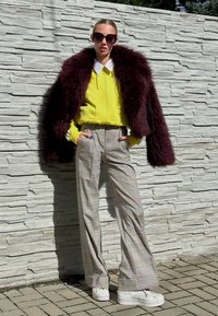 Casaco de pelo sintético bordô sobre um hoodie amarelo brilhante; calças largas estampadas em bege e cinza; ténis brancos; óculos de sol grandes.