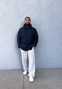 Μαύρο μπουφάν με ψηλό γιακά, φορεμένο πάνω από άσπρα φαρδιά παντελόνια και ανοιχτόχρωμα παπούτσια. Λείο ύφασμα και ραφές με πλέγμα.