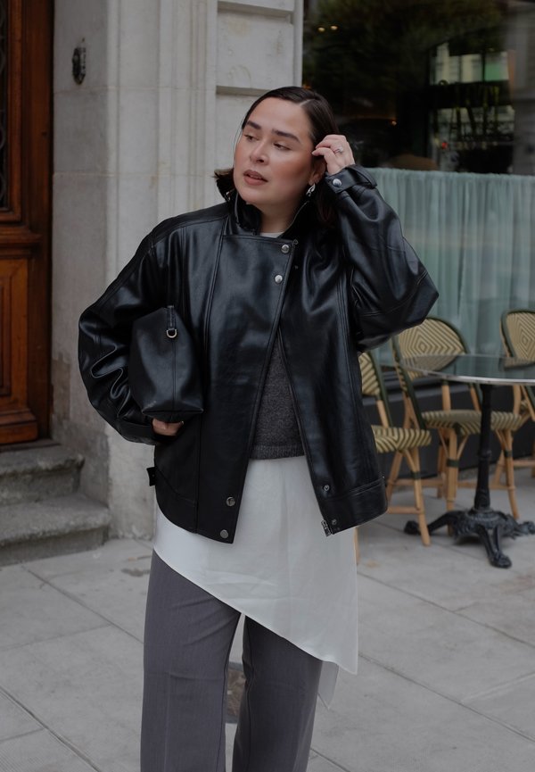 Femme portant une veste en cuir noire, un haut blanc asymétrique et un pantalon gris, debout devant un café avec des chaises en osier et une table en verre.