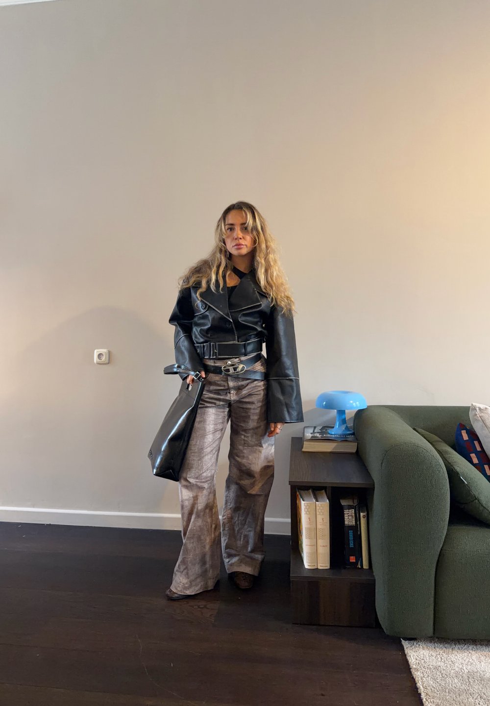 Femme aux cheveux longs portant une veste en cuir noire et un pantalon ample à motifs, debout à côté d'une table d'appoint en bois foncé et d'un canapé vert dans une pièce.