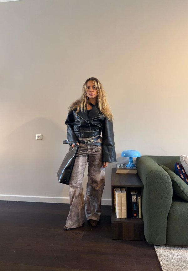 Femme aux cheveux longs portant une veste en cuir noire et un pantalon ample à motifs, debout à côté d'une table d'appoint en bois foncé et d'un canapé vert dans une pièce.