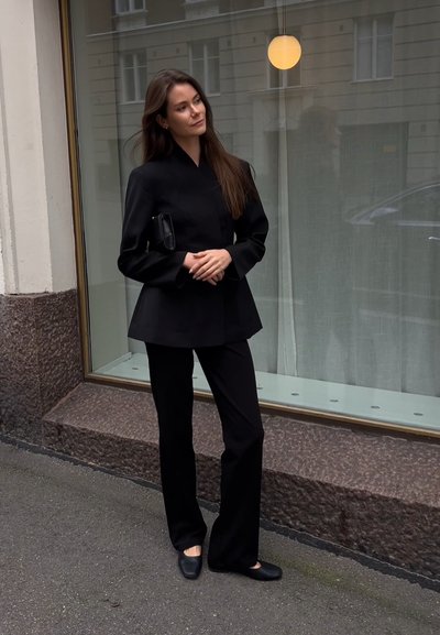 Chaqueta negra ajustada con cuello alto, combinada con pantalones anchos negros. Diseño elegante, detalles mínimos y textura de tela suave.