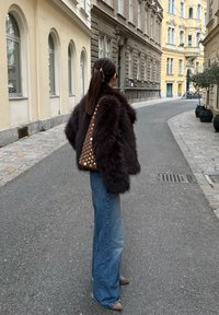 Veste en fausse fourrure marron à manches larges, associée à un jean évasé bleu et des talons beiges. Grand sac marron avec découpes circulaires. Cadre urbain en extérieur.