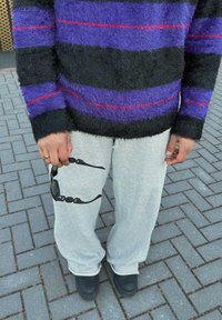 Černý a fialový chlupatý svetr s červenými pruhy, šedé tepláky, držící černé sluneční brýle, s černou obuví na vydlážděném povrchu.
