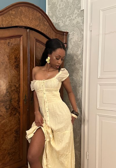 Mujer con vestido crema de botones y hombros descubiertos posa junto a un armario de madera, lleva grandes pendientes de flores y pulseras gruesas en una habitación decorada.