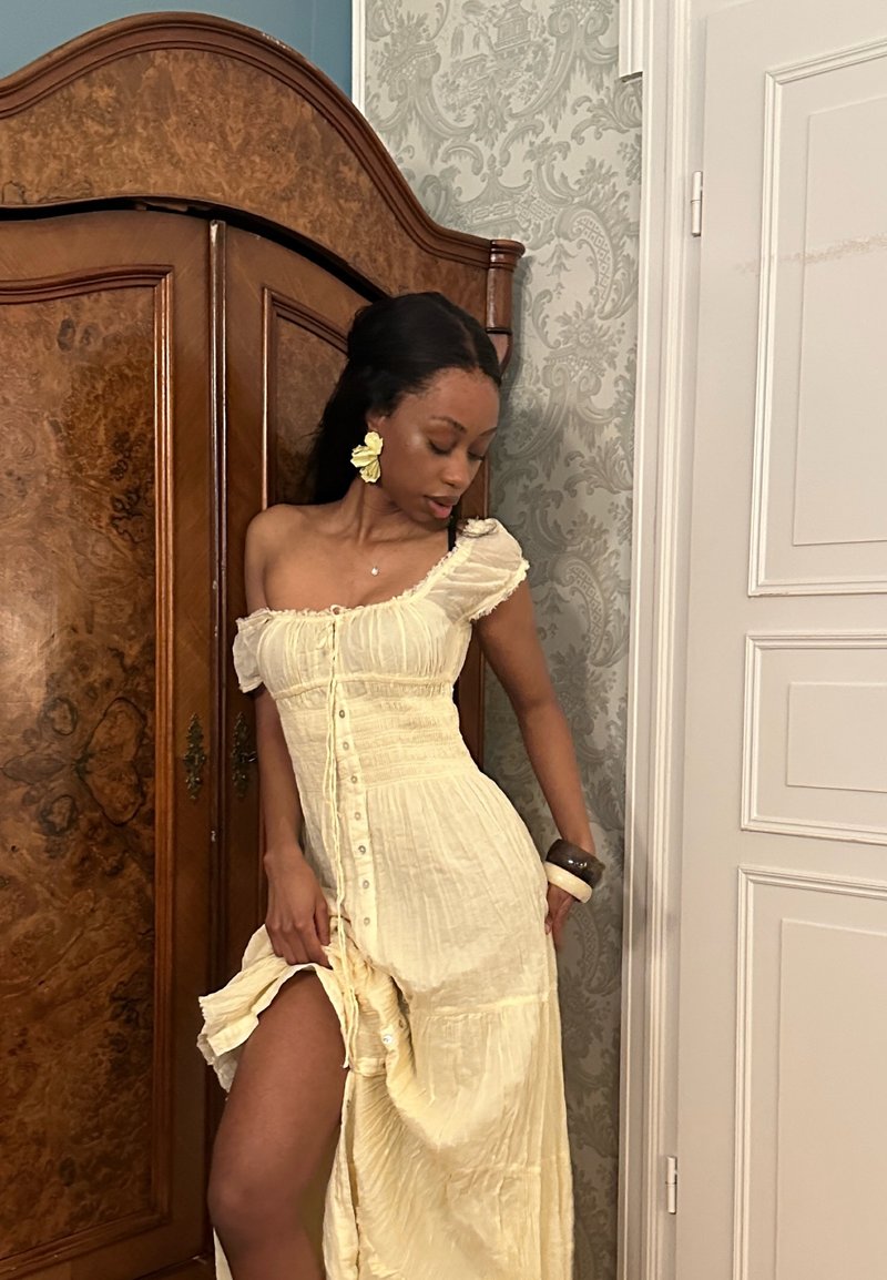 Kvinde iført en cremefarvet, skulderfri kjole med knapper poserer ved siden af et træskab. Hun bærer store blomsterøreringe og chunky armbånd i et udsmykket rum.