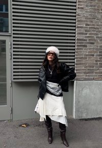 Schwarze Lederjacke, weißer Lagenrock, Kunstpelzmütze, übergroße Sonnenbrille, kunstvoller weißer Schal, braune kniehohe Stiefel, lässige Haltung.