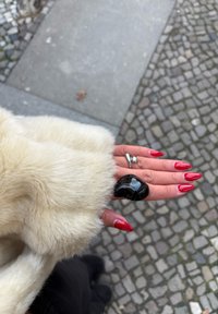 Kabát z umelého kožušina s jemnou béžovou textúrou; ruka s lesklými červenými nechty nosiaca čierny masívny prsteň. V pozadí je viditeľné dlažobné kamene.