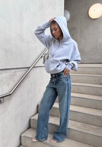 Harmaa vetoketjuhuppari, jossa on rento leikkaus, yhdistettynä leveälahkeisiin sinisiin farkkuihin. Seisoo portailla, pitää päätä kiinni, pienessä laukussa.