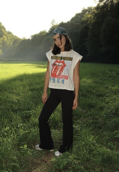 Camiseta blanca con gráfico de los Rolling Stones y logo de lengua roja, combinada con pantalones acampanados negros y zapatillas plateadas, en un campo de hierba.