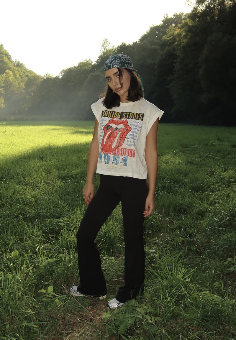 Camiseta branca com gráfico dos Rolling Stones e logo da língua vermelha, combinada com calças pretas com boca de sino e ténis prateados, situada num campo gramado.