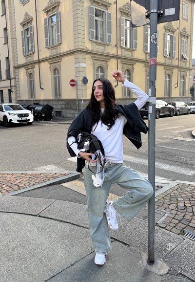 Camisa blanca de manga larga, chaqueta negra sobre los hombros, jeans holgados en azul claro, zapatillas blancas, llevando un bolso negro con accesorios.