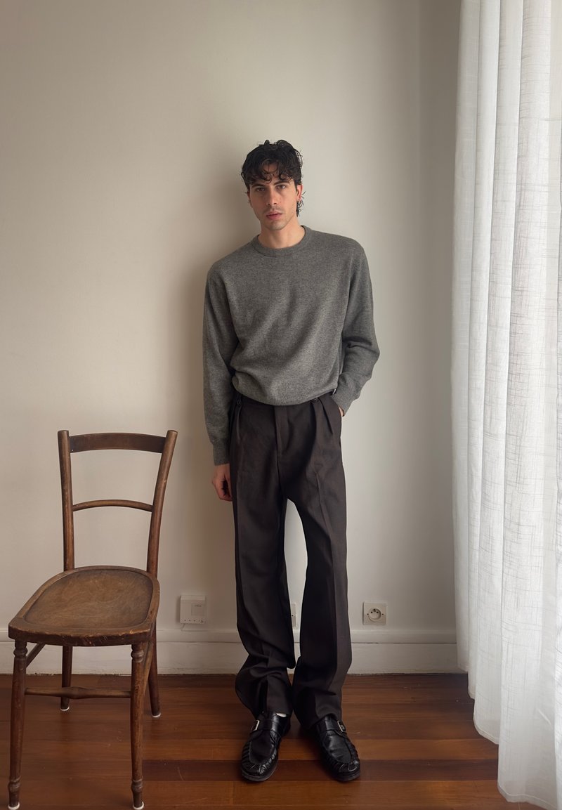 Young man standing indoors near a wooden chair and white curtains, wearing a gray sweater, black trousers, and black shoes.