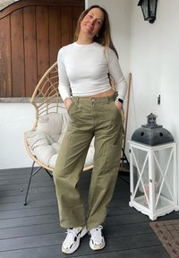 Ελαιόχρωμο cargo παντελόνι με πλαϊνές τσέπες, συνδυασμένο με ένα εφαρμοστό άσπρο μακρυμάνικο μπλούζα και άσπρα αθλητικά παπούτσια, που στέκει σε μια ξύλινη πλατφόρμα.