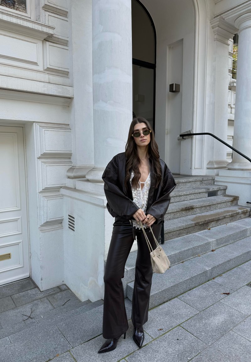 Chaqueta negra de gran tamaño sobre una blusa blanca con detalles de perlas, combinada con pantalones de cuero negros acampanados y tacones de punta. Bolso beige.