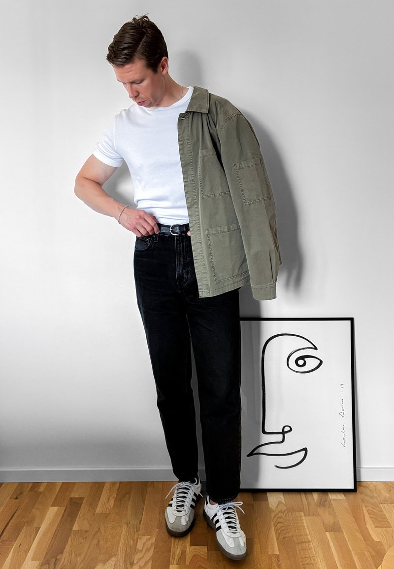 Homme portant un t-shirt blanc, un jean noir et des baskets ajuste sa ceinture avec une veste verte posée sur l'épaule à côté d'une œuvre encadrée au visage minimaliste.