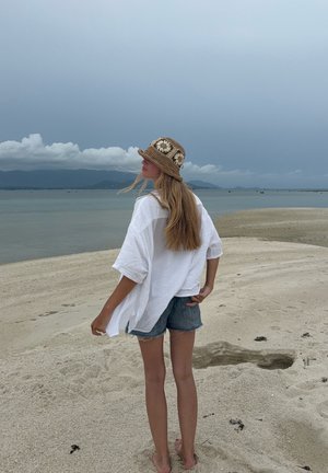 Camisa blanca, ligera y texturizada; pantalones cortos de mezclilla; sombrero de paja marrón con acentos florales. Playa de arena y agua de fondo. Cielo nublado.