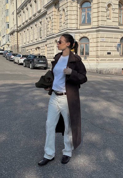 Mujer vestida con abrigo largo oscuro, camiseta blanca, pantalones blancos, gafas de sol, sosteniendo una chaqueta doblada, de pie en la calle cerca de un edificio de piedra beige.
