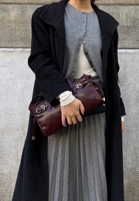 Person in einem schwarzen Mantel und einem grauen Cardigan, der eine dunkelburgunderfarbene Lederhandtasche vor einer strukturierten grauen Wand hält.