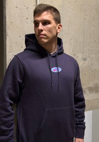 Marineblå hettegenser laget av mykt stoff, med en frontlomme og en rund Nike-logo med rød skrift på blå bakgrunn.