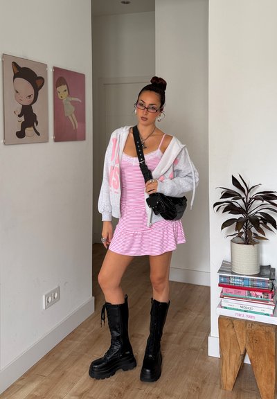Joven mujer con vestido rosa a rayas, sudadera gris con capucha, botas negras y gafas, de pie en el interior cerca de dos obras de arte en la pared y una planta sobre libros apilados.